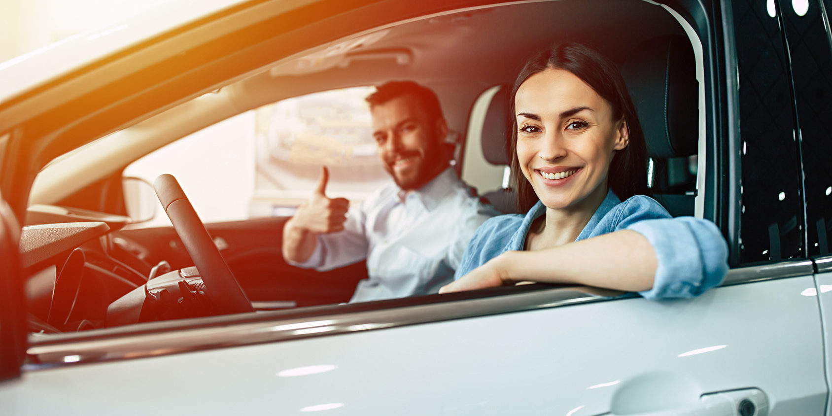 Happy smiling young couple chooses and buying a new car for the family in the dealership in sunlight.